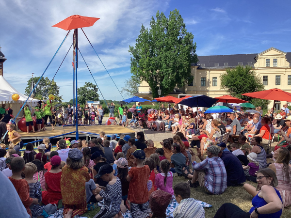 Kinder und Eltern unter bunten Sonnenschirmen sitzen im Kreis um eine Bühne
